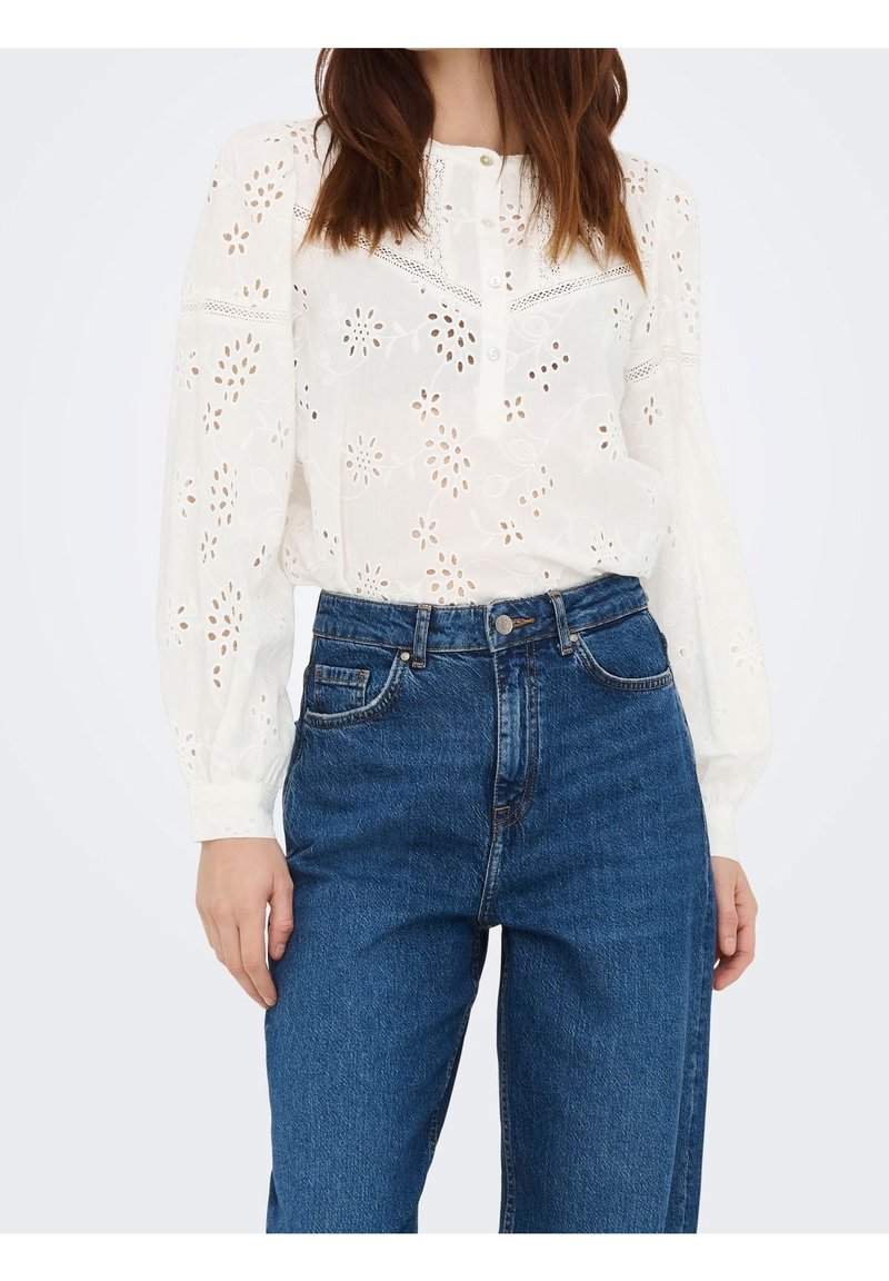 Blouse blanche à manches longues avec broderie florale à jour, associée à un jean en denim bleu taille haute.