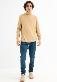 Suéter de cuello alto beige, de tejido ligero, diseño ajustado; combinado con vaqueros azules y zapatillas beige. Look simple y casual con una textura suave.