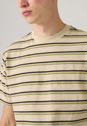 Jeune homme portant un t-shirt beige avec des rayures horizontales vert olive et marron foncé, ainsi qu'un petit logo blanc Levi's sur la poitrine.