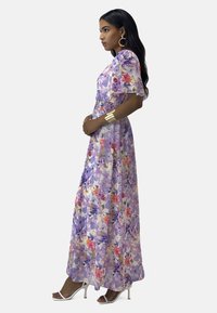 Maxi-jurk van doorschijnende paarse stof met bloemenpatronen. Heeft korte fladdermouwen, een gerimpelde taille en een split aan de zijkant. Draagt zilveren hakken.