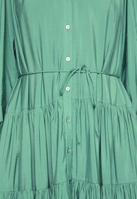 Robe verte boutonnée à la taille froncée, fine ceinture nouée à l'avant, et détails plissés sur la jupe. Pas de personnes ni d'arrière-plan visibles.