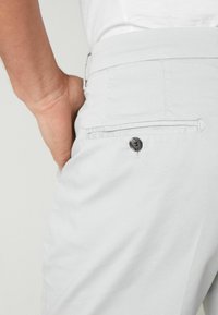 Pantaloni grigi chiari con una tasca posteriore con chiusura a bottone. Texture liscia e vestibilità sagomata. Dettaglio di cucitura sottile lungo la cintura.