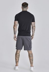Schwarzes Sport-T-Shirt mit kurzen Ärmeln, kombiniert mit grauen Cargo-Shorts mit Taschen, an einer Person mit Tattoos, vor einem neutralen Hintergrund.