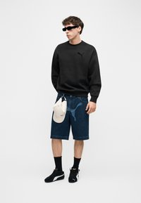 Νεαρός άνδρας που φοράει μαύρο φούτερ, σκούρες τζιν βερμούδες με το λογότυπο της Puma, μαύρα παπούτσια Puma, μαύρες κάλτσες, γυαλιά ηλίου και κρατάει ένα λευκό καπέλο.