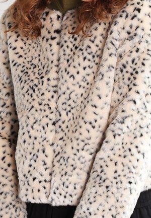 Personne aux cheveux bouclés portant un manteau en fausse fourrure blanche avec des taches noires denses et un col montant sur un haut vert.