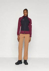 Γυναίκα που στέκεται με ένα μπορντώ και μαύρο φούτερ με φερμουάρ, τατανίσιο ίσιο παντελόνι και μαύρες ορειβατικές μπότες σε απλό φόντο.