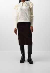 Maglione lavorato a maglia crema con motivo texturizzato e maniche a sbuffo, abbinato a una gonna midi in corduroy marrone scuro e stivali neri al ginocchio con suola spessa.