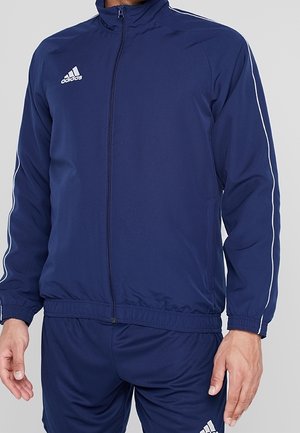 Mężczyzna w granatowym dresie Adidas z białymi paskami na rękawach oraz logo Adidas na piersiach i udzie.
