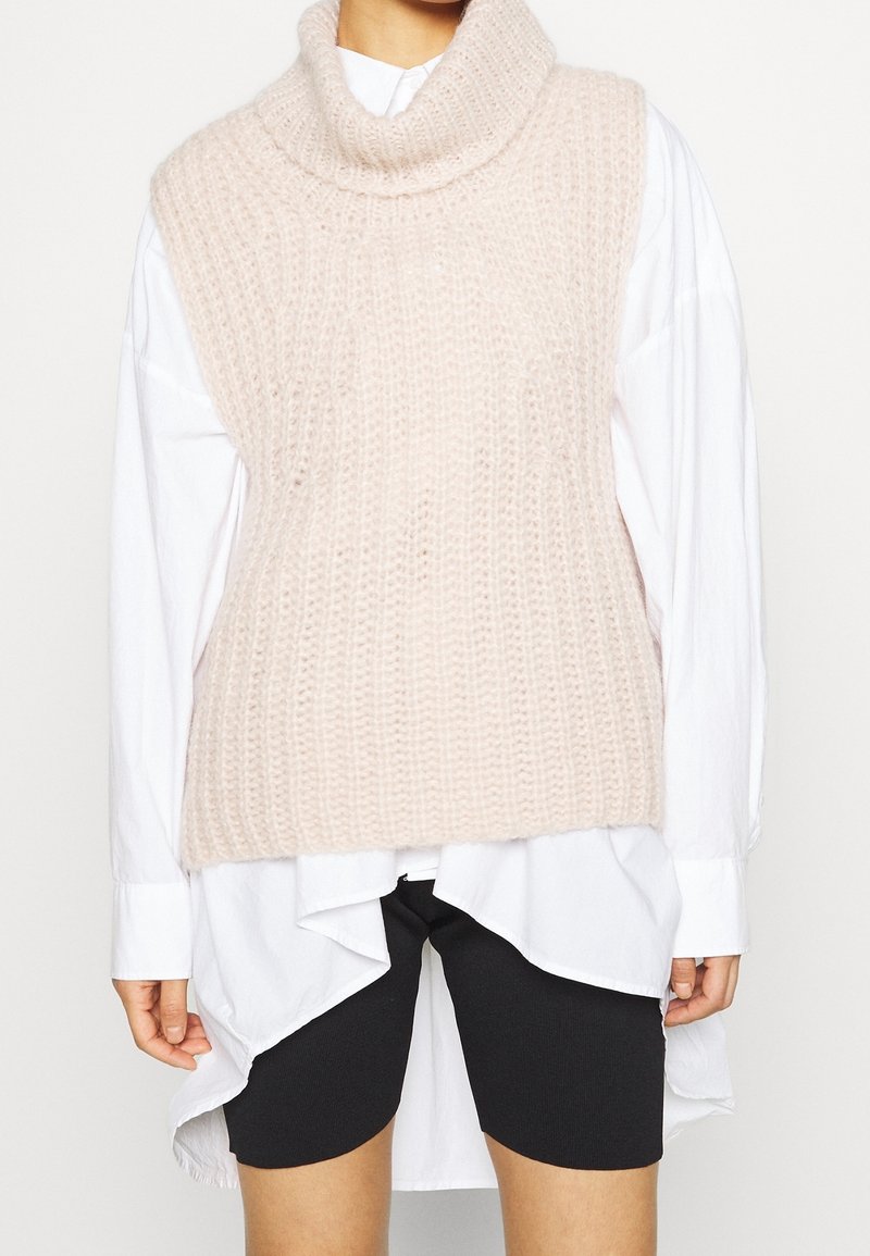Chunky, gestrickter beiger ärmelloser Rollkragenpullover über einem weißen Hemd und schwarzen Shorts. Der Stoff hat eine weiche Textur mit einem gerippten Muster.