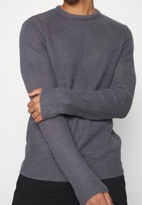 Osoba w teksturowanym szarym swetrze z prążkowanymi mankietami i dołem, krzyżująca jedną rękę na drugiej, na jednolitym tle.