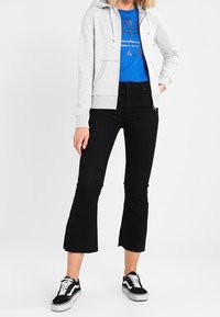 Sudadera gris claro con bolsillos frontales, combinada con una camiseta gráfica azul, pantalones de campana negros con canalé y zapatillas negras con detalles blancos.