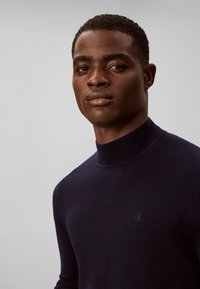 Ung sort mand iført en mørk rullekravetrøje, der ser selvsikkert på kameraet mod en ensartet lys baggrund.