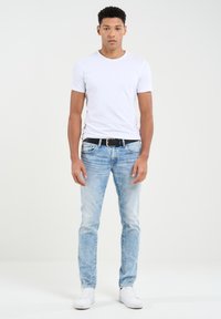 Baltas apdailos marškinėliai, šviesiai mėlynos džinsinės kelnės su išblukusiu efektu, juodas diržas ir balti sportbačiai, stovintys priešais vientisą foną.