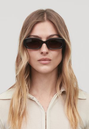 Lunettes de soleil noires, rectangulaires, avec des montures épaisses et des verres sombres, présentées sur un mannequin avec de longs cheveux ondulés et un haut couleur crème.