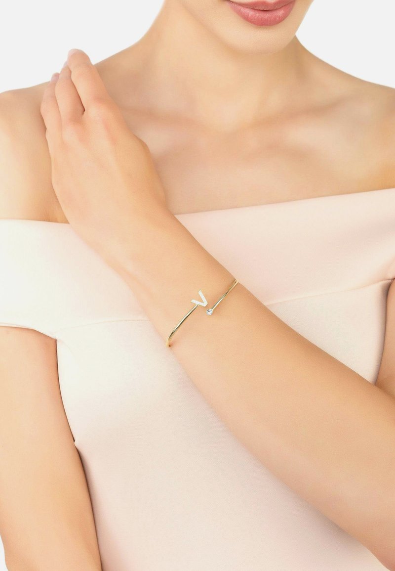 Aranyszínű karkötő fehér "V" díszítéssel és egy kis átlátszó kővel, amely egy csupasz karon fekszik egy semleges h�áttér előtt.