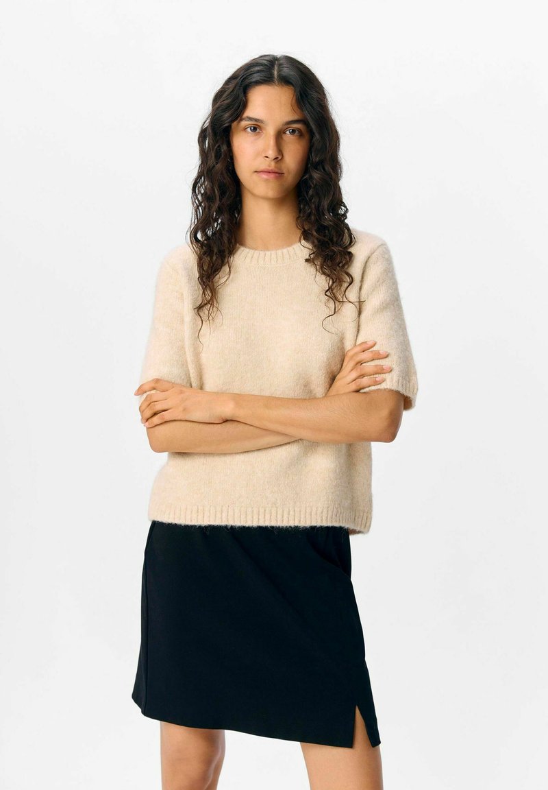 Kortsleeve genser i myk beige stoff med ribbestrikket hals, kombinert med et svart miniskjørt med sidespalter.