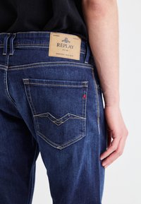 Mörkblå denimjeans med en läderetikett som säger "REPLAY." Har gula sömmar och en distinkt okdesign på bakfickorna.