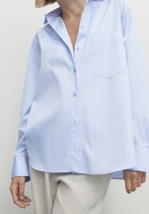 Osoba ubrana w luźną jasnoniebieską koszulę na guziki z kieszenią, w połączeniu z jasnymi spodniami, na jednolitym tle.