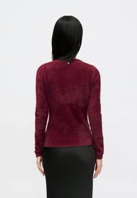 Egyenes fekete hajú nő egy testhez simuló burgundi színű bolyhos pulóverben és egy fekete szoknyában áll egy sima fehér háttér előtt.