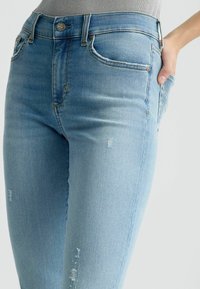 Heled sinine teksapüksid, mis on slim fit stiilis, millel on õrn kulumine, viie tasku disain ja klassikaline nööpkinnitus vööl.