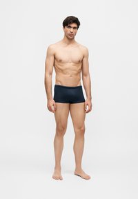 Ung mann står barbeint iført mørk blå boxer mot en ensfarget lys bakgrunn.