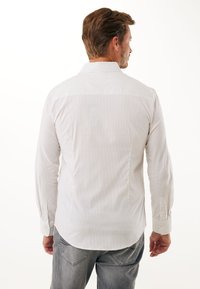 Camisa de botones blanca a rayas con mangas largas, que presenta un cuello estándar y un corte recto, combinada con jeans grises.