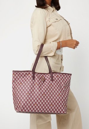 Vrouw in beige jas en broek met een grote bordeauxrode en witte geruite draagtas met dubbele handvatten.