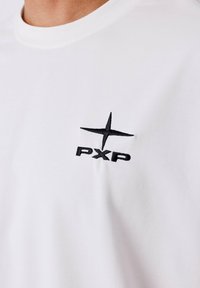 Camiseta blanca de manga corta con un logo bordado en negro "PXP" con un diseño de estrella en el área del lado izquierdo del pecho. Fabricada en suave algodón.