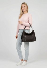 Moteris dėvi rožinį megztinį ir šviesius džinsus, laiko didelę juodą odinę rankinę su priekinėje kišenėje užtrauktuku, šypsosi į kamerą.