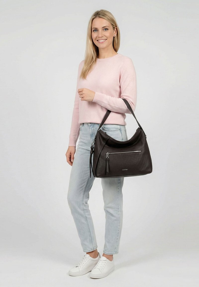 Moteris dėvi rožinį megztinį ir šviesius džinsus, laiko didelę juodą odinę rankinę su priekinėje kišenėje užtrauktuku, šypsosi į kamerą.