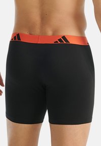 Svarta boxer-kalsonger med en enfärgad design, med ett orange elastiskt midjeband prytt med svarta Adidas-ränder. Slät textur.