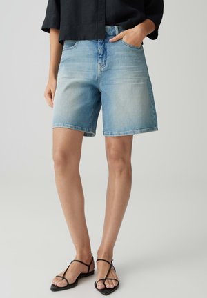 Femme portant un short en denim bleu clair, un haut noir et des sandales plates noires à brides, debout avec une main dans la poche.