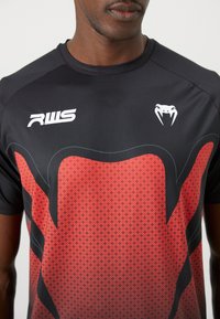 Schwarz-rote Sportshirt mit Diamantmuster, kurzen Ärmeln und flachem Kragen. Mit Logo auf der Brust in Weiß. Glatte Stoffoberfläche.