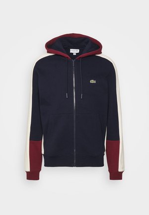 Marineblauwe hoodie met rits, bordeaux kap en manchetten, witte strepen op de mouwen, voorzakken en klein groen krokodillenlogo op de borst.