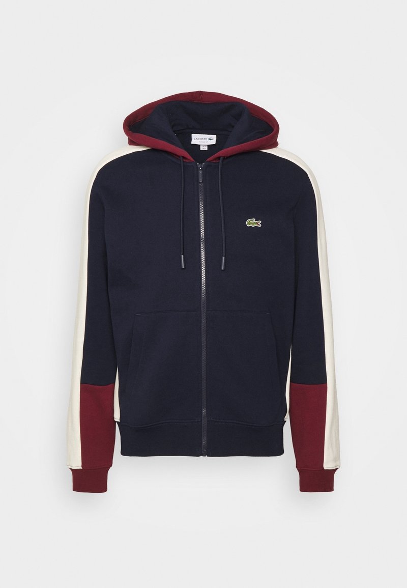 Sweat à capuche zippé bleu marine avec capuche et poignets bordeaux, bandes blanches sur les manches, poches avant et petit logo crocodile vert sur la poitrine.