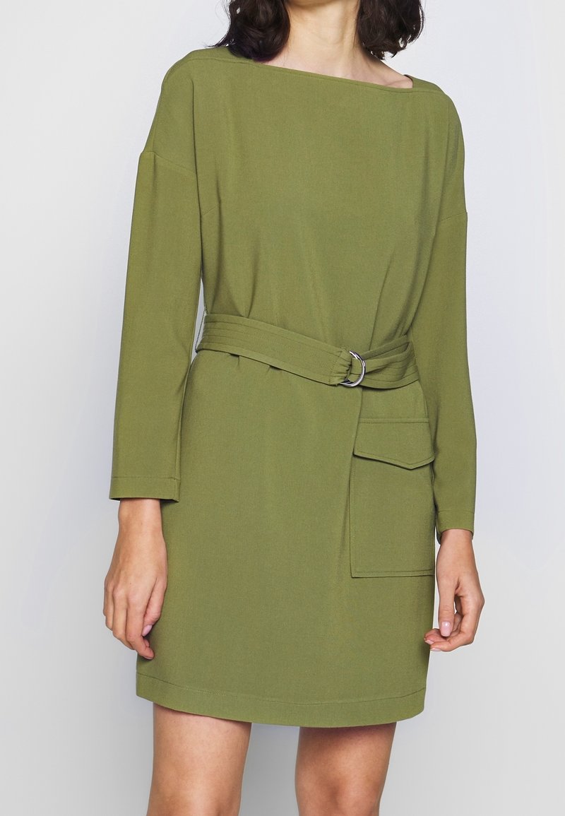 Olivgrön klänning med boxig form, långa ärmar och bältad midja. Inkluderar sidofickor och en mjuk tygliknande textur.