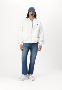 Chaqueta bomber de forro polar blanca con cierre de cremallera, gorra de béisbol azul, jeans azules y zapatillas blancas. Logotipo notable en el pecho de la chaqueta.