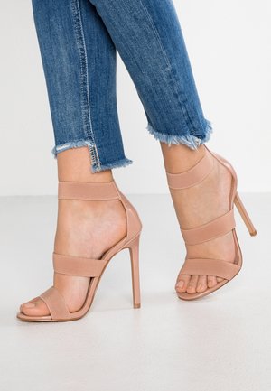 Nude højhælede sandaler med tre elastiske stropper. Glat tekstur, åbent tådesign og en slank stilethæl. Båret med slidte jeans.