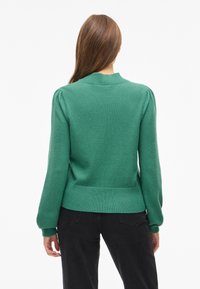 Grön stickad tröja med ribbad krage och midjeband, har puffärmar och en mjuk textur, bärs med mörka byxor.