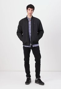 Black quilted bomber jacket with ribbed cuffs, worn over a light blue plaid shirt, paired with black pants and black sneakers.