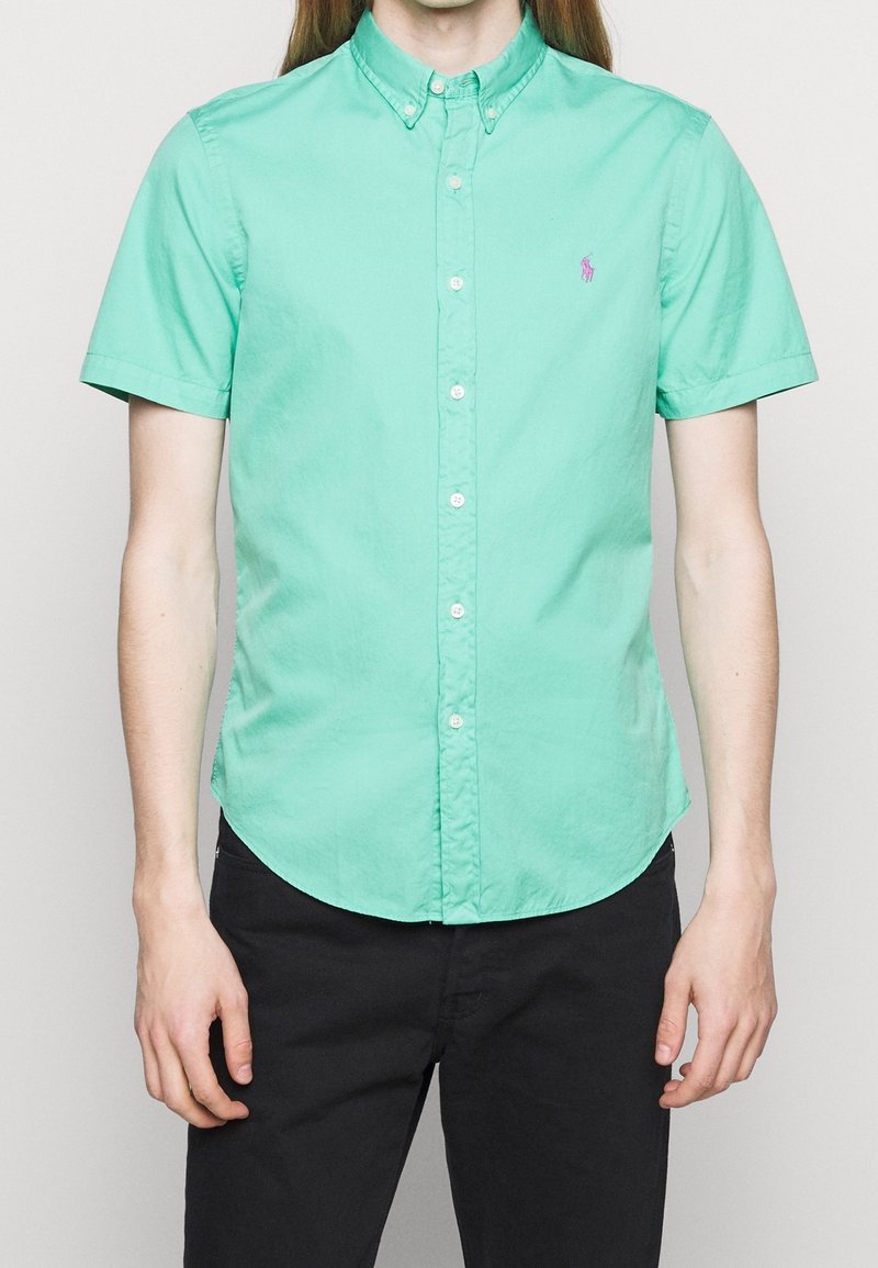 Košile s krátkým rukávem v mátově zelené barvě ze 100% bavlny, s kulatým lemem a decentním fialovým logem na hrudi. Klasický límec a bílé knoflíky.