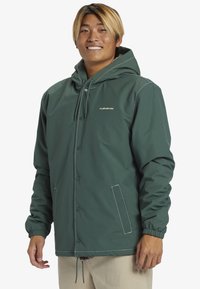 Veste verte avec capuche, fabriquée en matériau imperméable. Elle possède des boutons-pression, des poignets élastiques et des poches avant avec des accents de couture blanche.