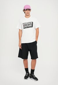 Balta T-krekls ar melnu "VERSACE COUTURE" druku, apvienots ar melniem šortiem un melniem sporta apaviem. Modelis valkā rozā cepuri.