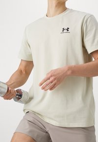 Ljusbeige bomullströja med rund hals, korta ärmar och en liten svart Under Armour-logotyp på bröstet, kombinerad med grå shorts.