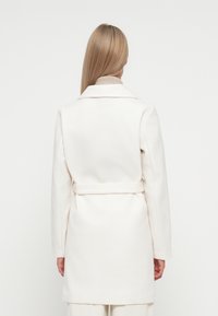 White, belted coat made of soft fabric with a structured design, featuring a classic collar and long sleeves. Plain texture.