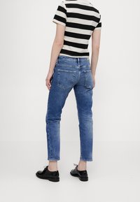 Osoba oblečená v modrých denimových džínách, černém a bílém pruhovaném krátkém rukávu a černých kožených šněrovacích botách, stojící na jednobarevném pozadí.