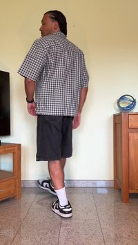 Camisa de manga curta, quadrada em preto e branco, combinada com calções de ganga pretos e sapatilhas pretas. A pessoa está de pé sobre um chão de azulejos com móveis por perto.