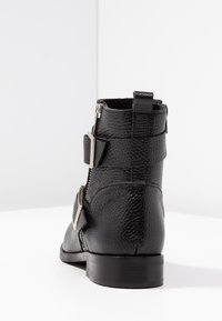 Botín negro de cuero hasta el tobillo con un acabado texturizado, que presenta dos hebillas ajustables y un zipper lateral para el cierre. Tacón plano.