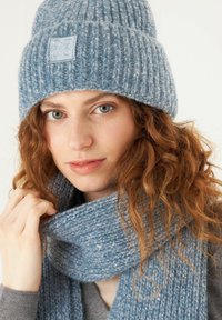 Light blue knitted beanie with a textured pattern and a small square logo; matching scarf in similar material draped around the neck.