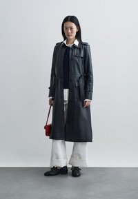 Długi czarny skórzany płaszcz typu trencz z guzikami, paskiem w talii i dużymi kieszeniami. W połączeniu z granatowym swetrem i szerokimi białymi spodniami.